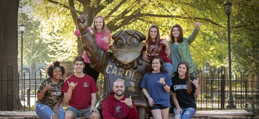 A group of diverse students surround the larger-than-life bronze Cocky statue, all flashing the spurs up hand sign.