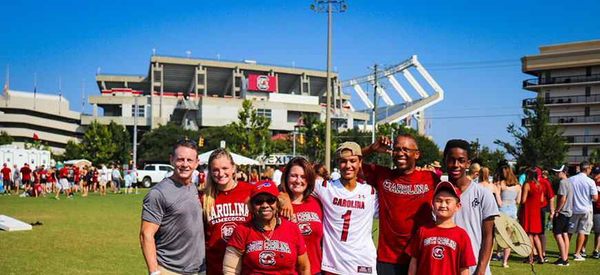 A large Gamecock family smiles brightly at the photographer while standing against the backdrop of Williams-Brice Stadium in the distance.