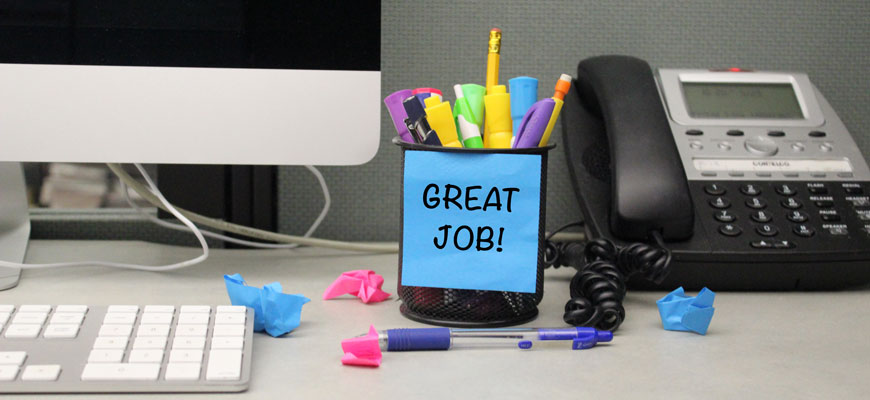 cup of pens on a desk with a phone and computer with post-it note that reads "great job"