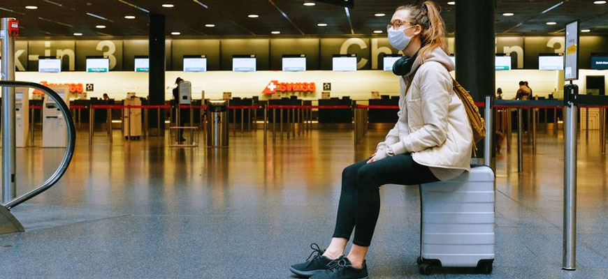 woman wearing a mask sits on a suitcase in an airport