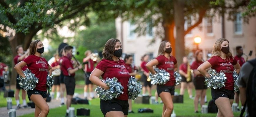 Carolina Coquettes dancing to the Carolina band on the horseshoe with pom poms. The coquettes are socially distant and wearing masks.