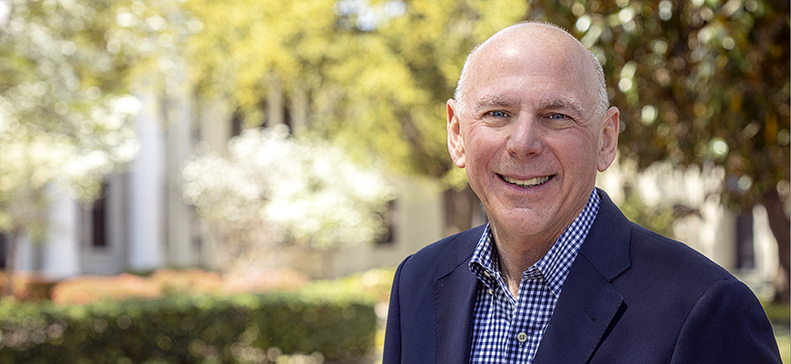 man in blue check shirt and blue jacket with trees and building in background