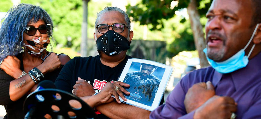 A woman and two men making the Wakanda forever signal. One man holds a photograph of Chadwick Boseman, who died from colon cancer at age 43.