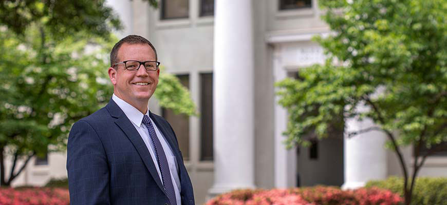 Tommy Hodges stands smiling in front of spring greenery and flowers and the white columns of Wardlaw College. 