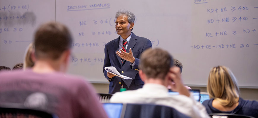 Professor Sanjay Ahire wearing blue jacket and tie in classroom with whiteboard and students in foreground