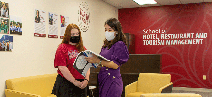 two women look at book in lobby of college of hospitality, retail and sport management