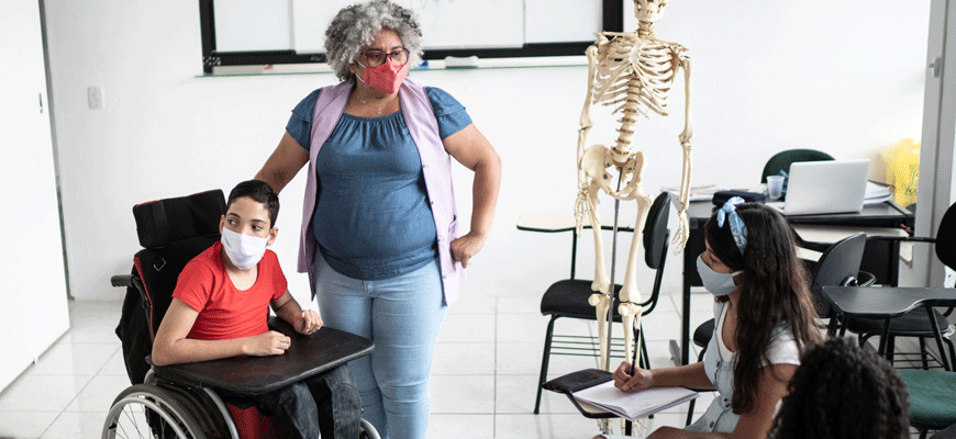 A teacher stands beside student using a wheelchair while other students sit in desks