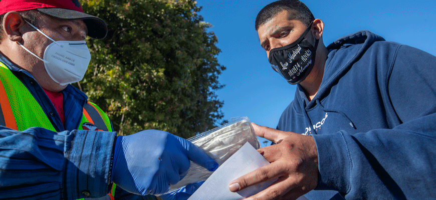 Man in red cap wearing gloves gives another man a packet of face masks