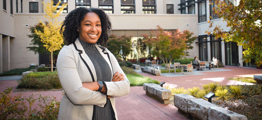 Glynnis Hagins wearing a black dress and cream and cream jacket stands in a brick courtyard with stone benches and fall trees in the background.