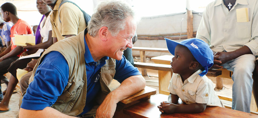 david beasley kneels and smile at a child wearing a blue ball cap