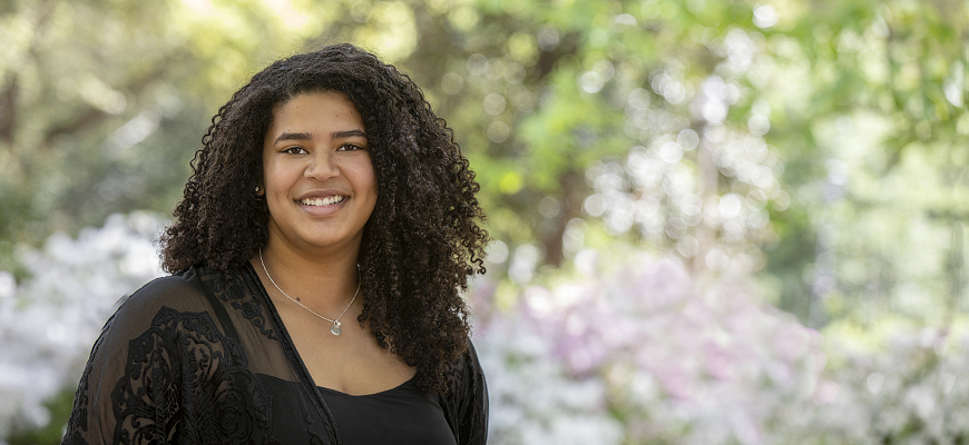 Graduating senior Brianna Lewis smiles as she stands to the left of the frame in front of a blurred outdoor background showing lush spring greenery and flowers.