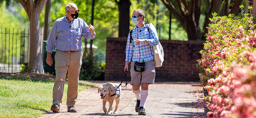 Journalist Harry Smith and Rhodes Scholar Jory Fleming walk along a brick path on the UofSC campus; flowers bloom on the right side of the photo. Fleming is walking his service dog, Daisy, as he talks with Smith.