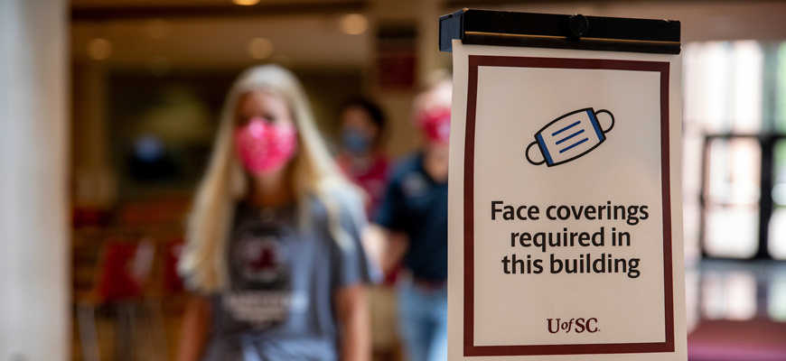 "Face coverings required in this building" sign in foreground, masked student in background.