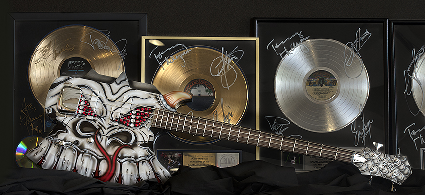 A Kiss bass painted with the face of a gray and red monster lays on its side in front of gold and platinum records.