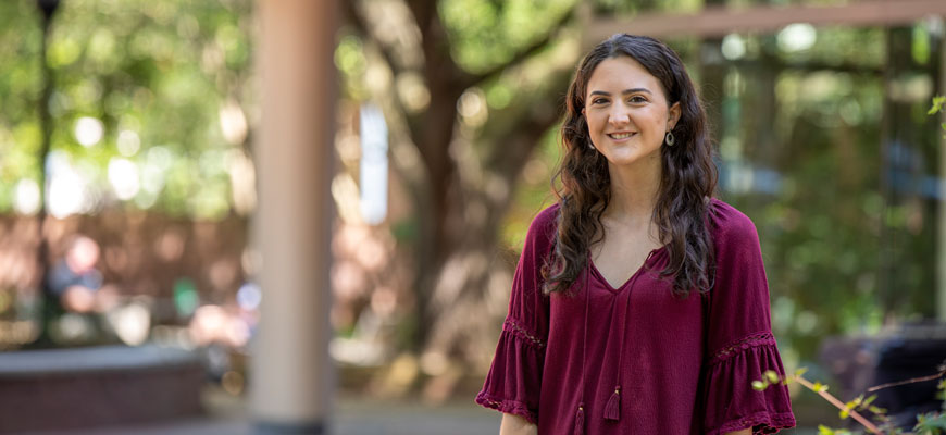 Biological sciences major Jessica Teresi stands to the right of the frame in focus; behind her in blurred background are trees, the Russell House patio and the Student Health Center. 