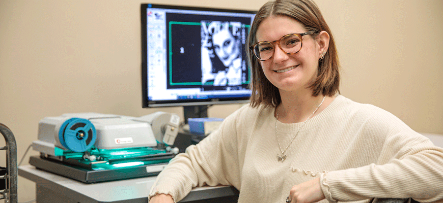 Student Kate Kuisel wearing glasses and tan top sits in front of a microfilm machine and computer screen