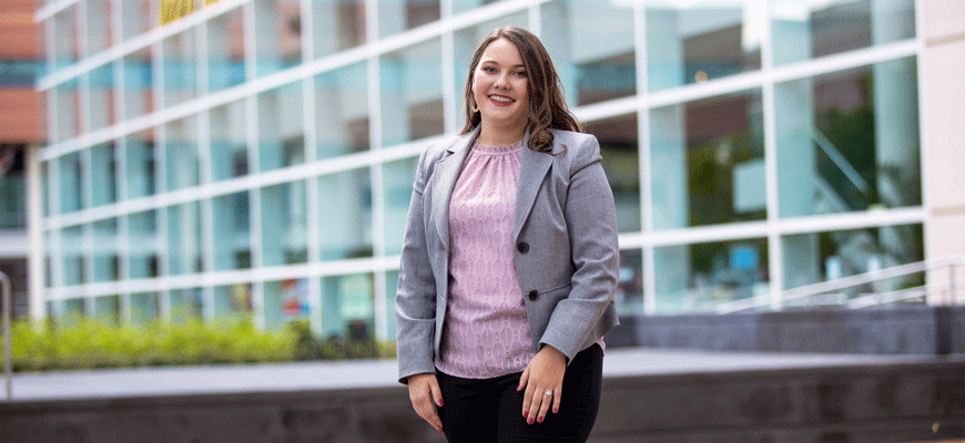 School of Music senior Madie Willard stands in front of the Koger Center wearing a gray jacket and pink top