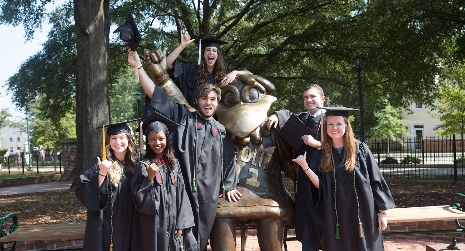 Students in commencement attire are gathered around the Cocky statue