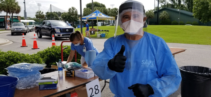Dr. Gerald Harmon in blue scrubs, face shield, mask and black gloves under a tent at a COVID testing site