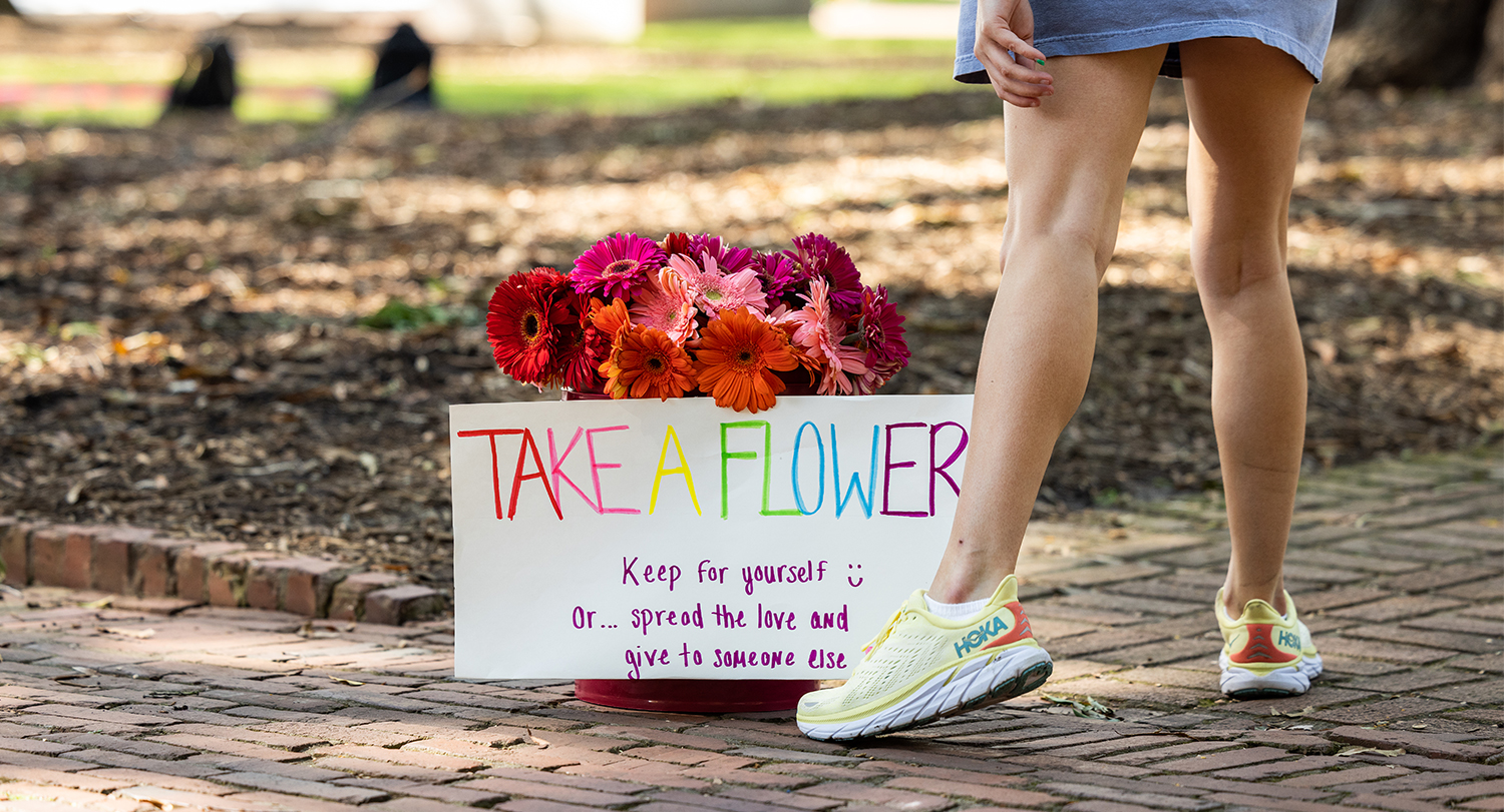 Student standing beside a bucket of flowers with a sign that says take a flower.