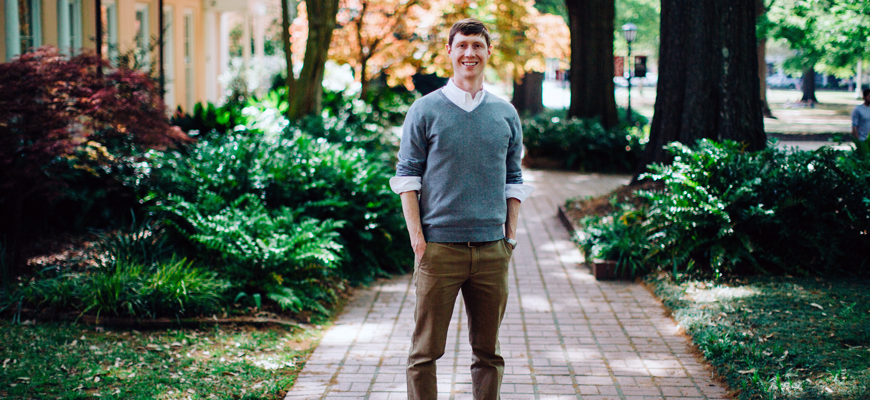 A man in a gray sweater stands on a brick pathway with trees in the background