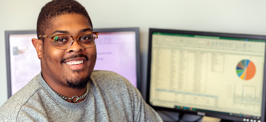 Man wearing glasses and a grey sweater sits in front of two computer screens