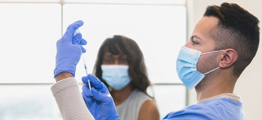 Doctor prepares syringe while woman in a face mask watches