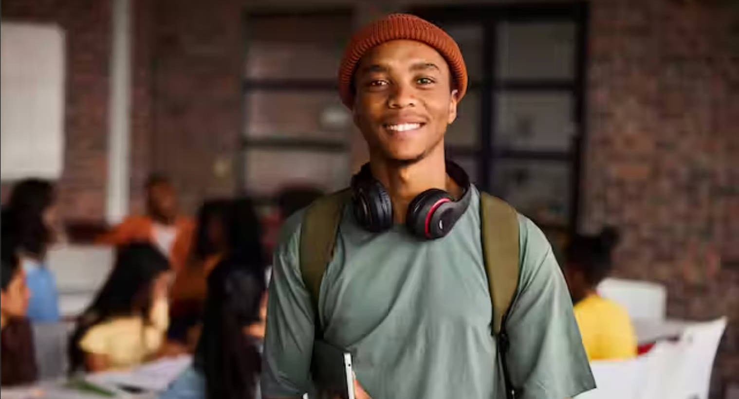 A man wearing a green shirt and orange hat with headphones around his neck smiles for the camera