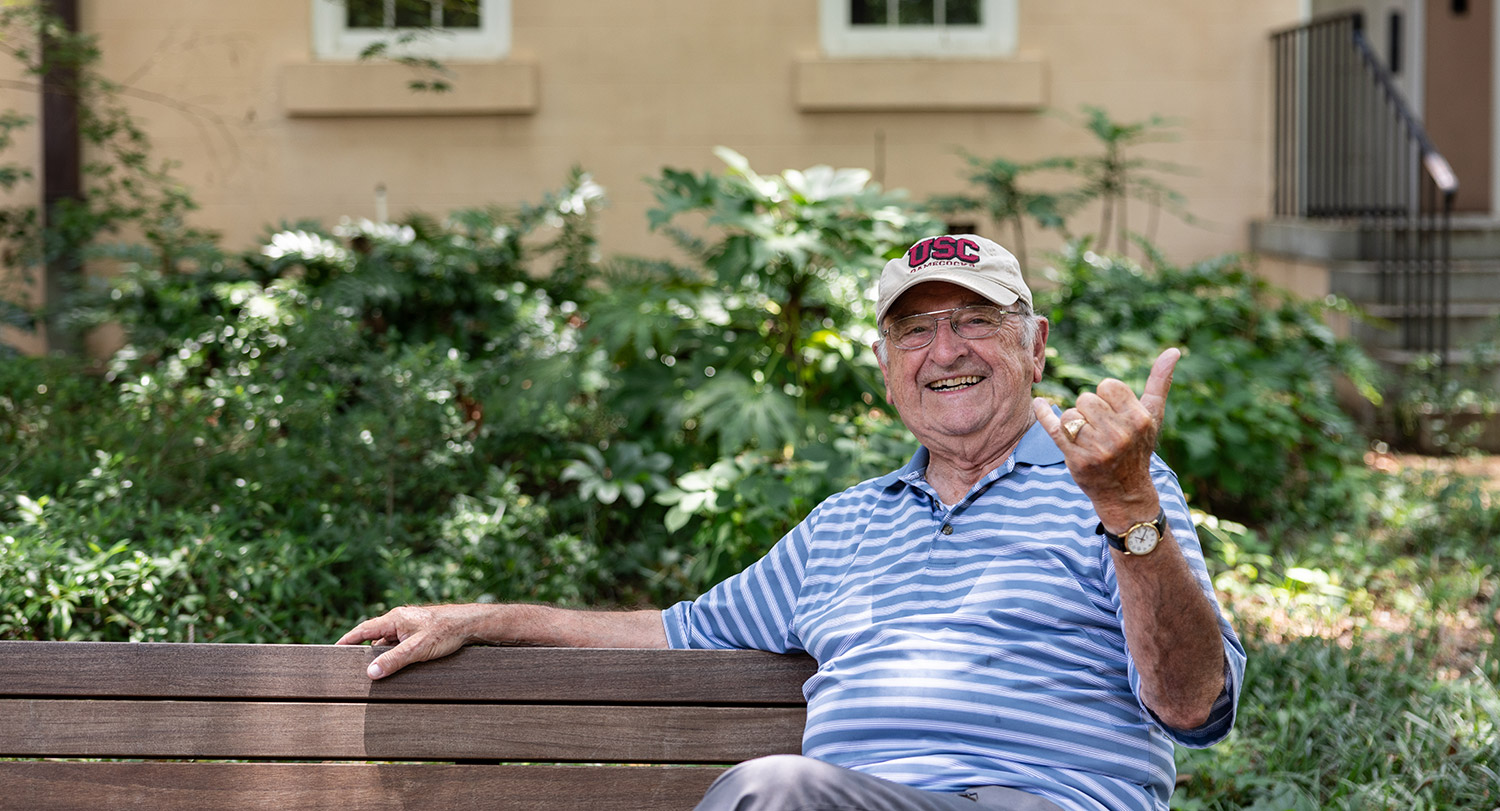 Stan Papajohn sitting on a Horseshoe bench wearing his USC hat.