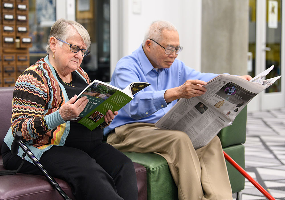 Two senior citizens in a library, reading a magazine and newspaper.