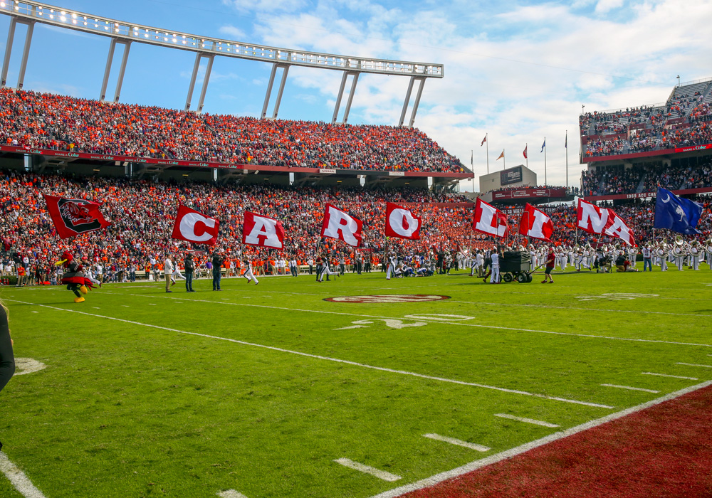 Cheerleaders run across a football field holding flags spelling out Carolina.