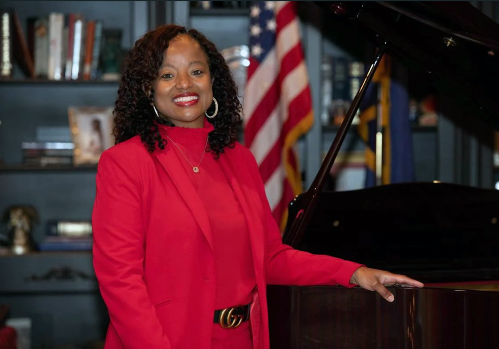 DeAndrea Benjamin stands next to a piano with a bookcase and a U.S. flag in the background