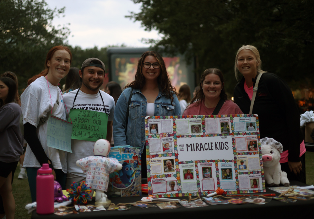 A man and four women stand behind a table with a sign that says, "Miracle Kids" surrounded by photos.