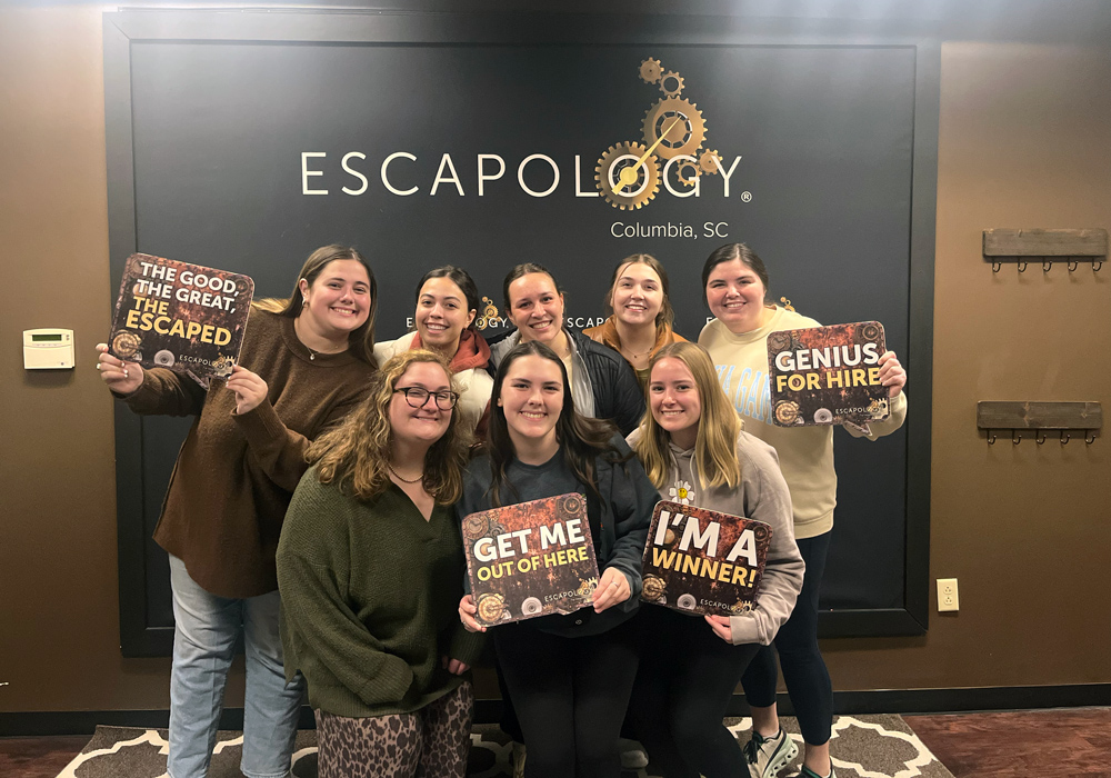 Eight girls stand in front of a wall that says, "Escapology Columbia, SC." Four girls hold small signs that read, "The Good. The Great. The Escaped," "Get me out of here," "I'm a winner!" and "Genius for hire."