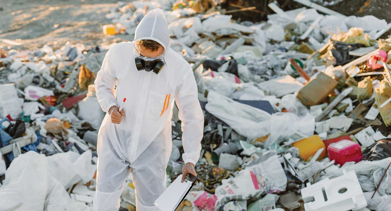 A person in protective gear with clipboard observing waste in a landfill.