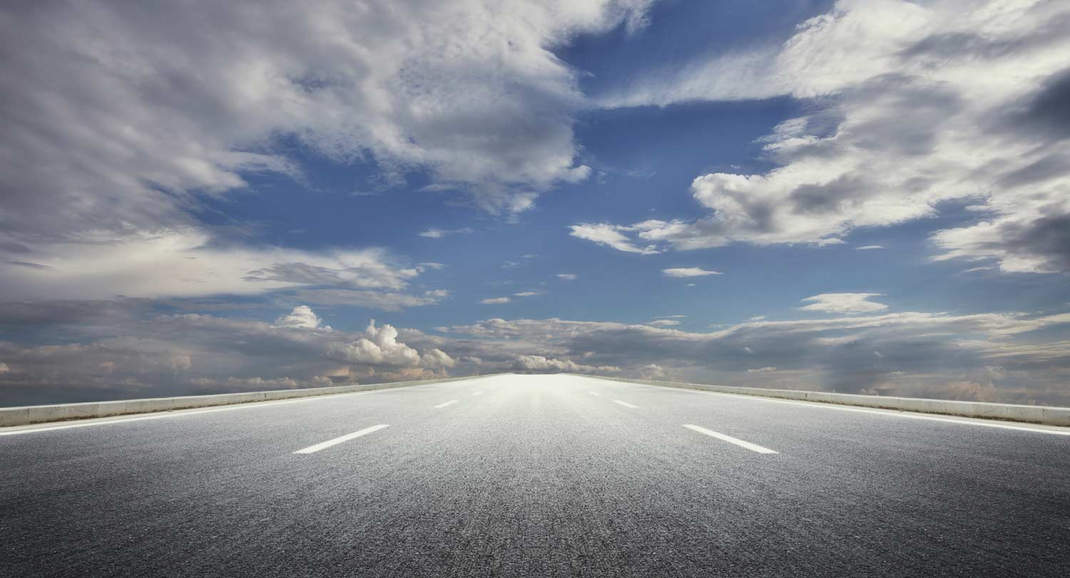 A view of a long, straight paved road with blue sky and clouds above.