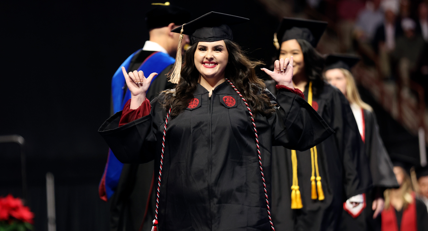 A student, wearing a graduation cap and gown, makes the hand signals for "Spurs up!" as she crossed the commencement stage