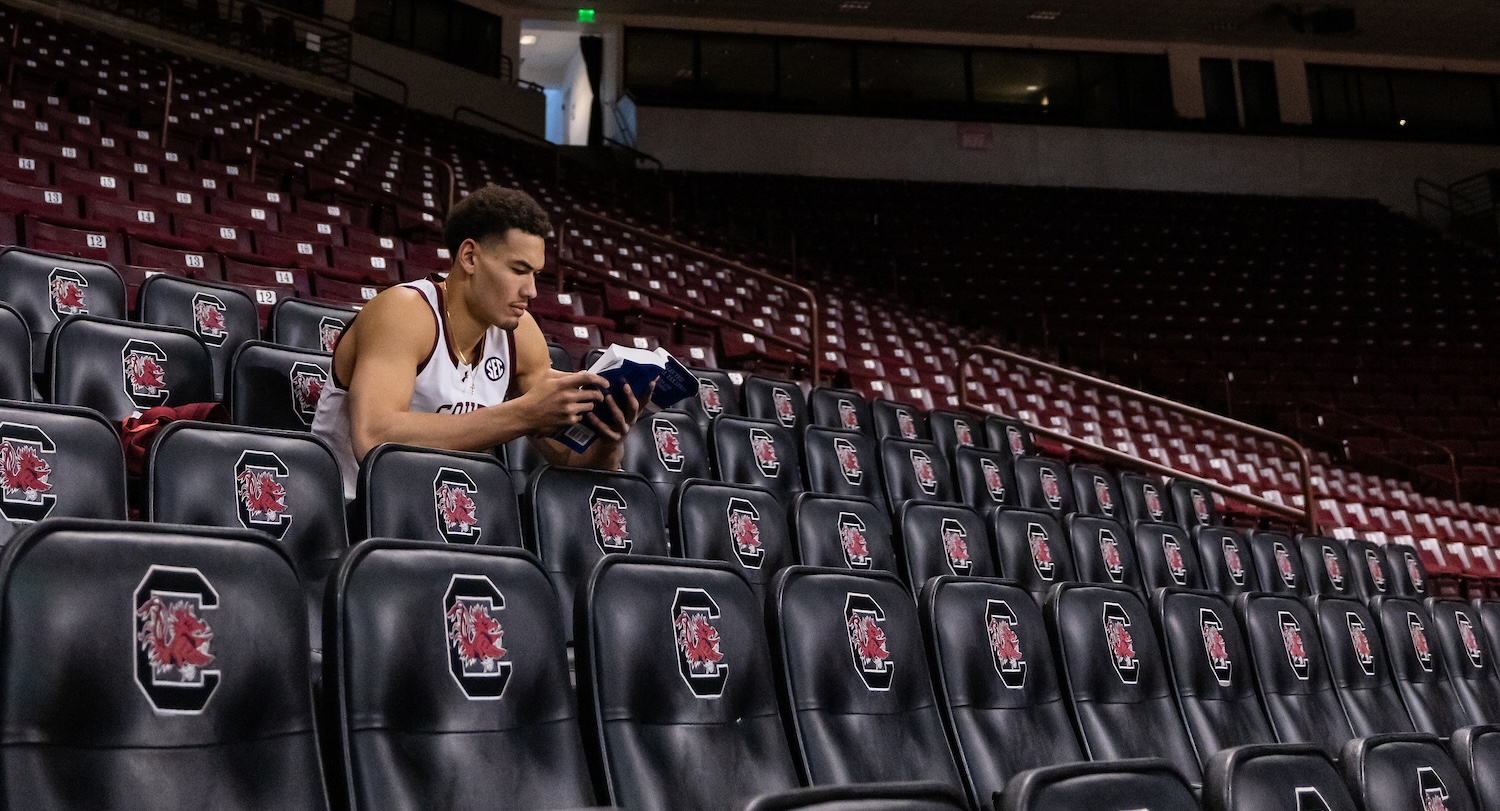 Man in white jersey with garnet South Carolina lettering reads blue textbook in empty basketball arena