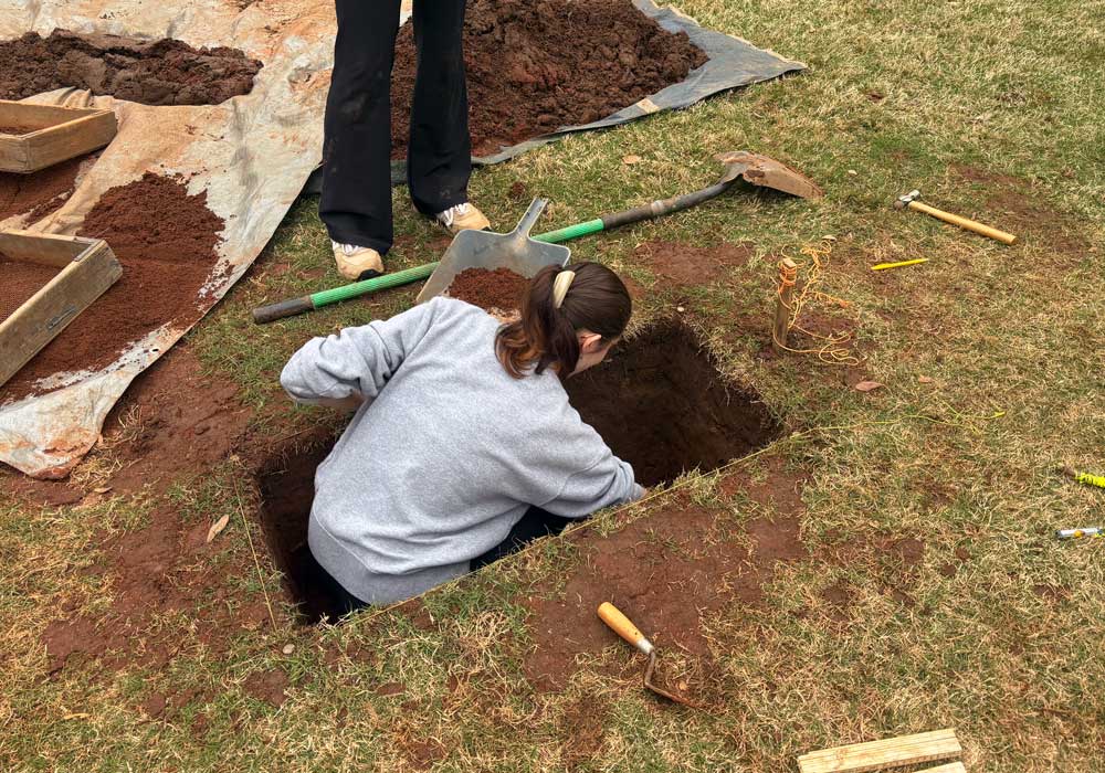 Student sits inside dug hole from archeological excavation
