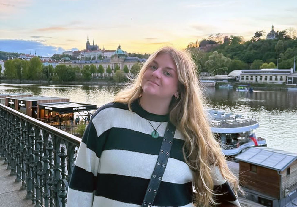 woman stands in front of a river with a large city in the background