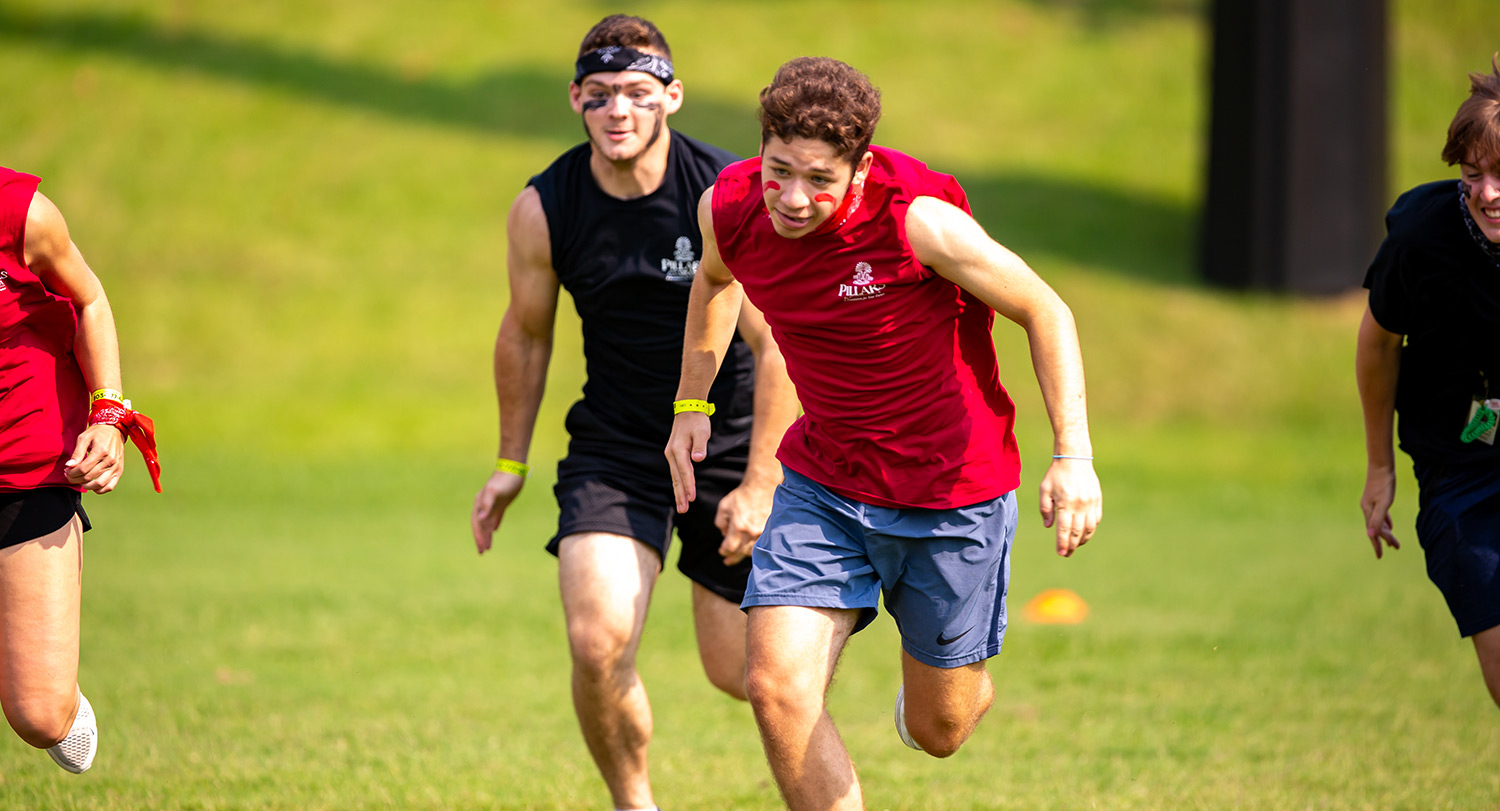 Two college-aged athletes run in competition in an open field in the heat.
