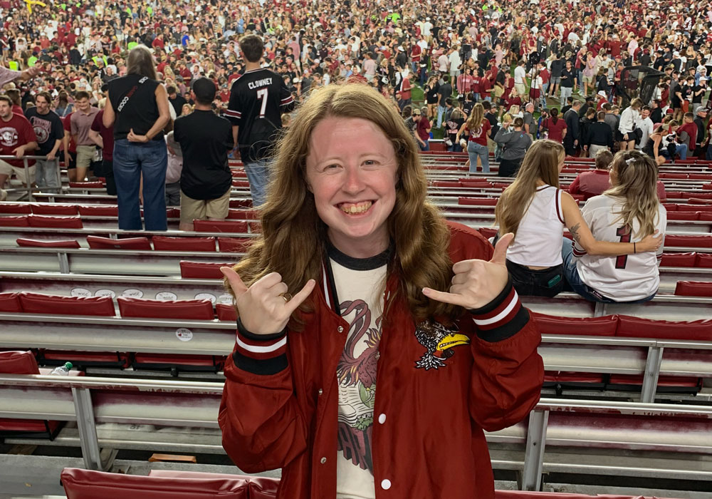 woman stands in a crowded football stadium giving the "spurs up" hand sign