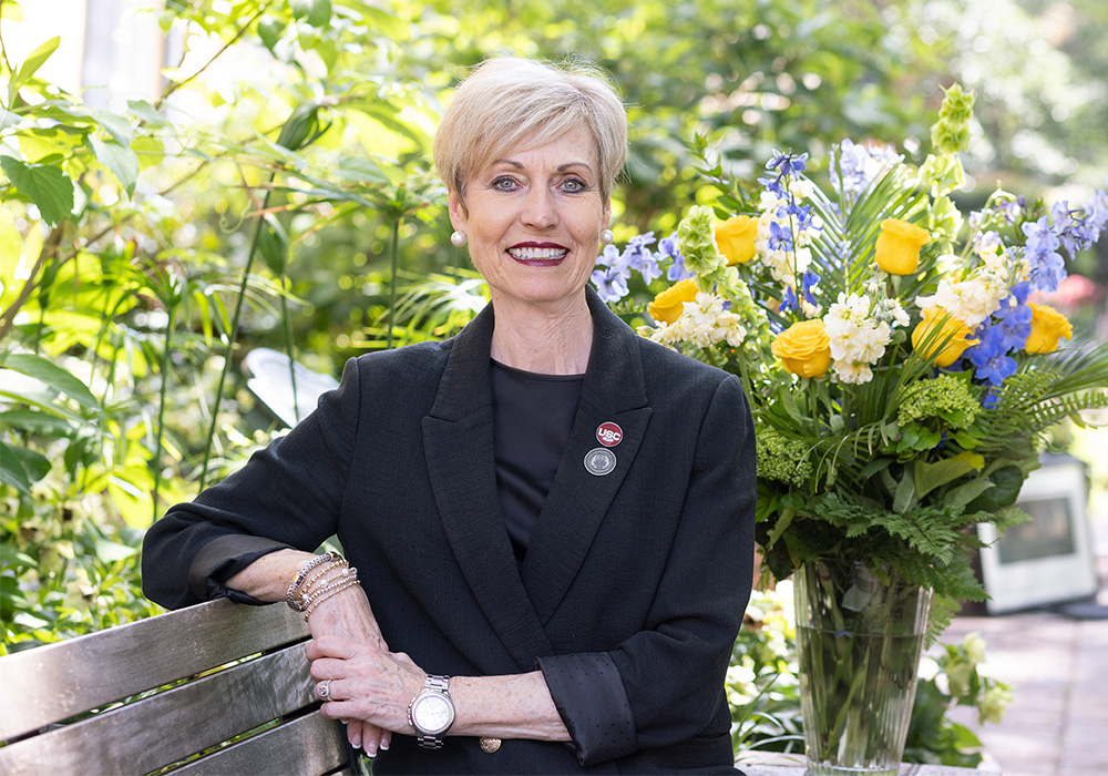 Susan Elkins sits on a bench on USC's historic Horseshoe with a vase of yellow, lavender and white flowers over her left shoulder.