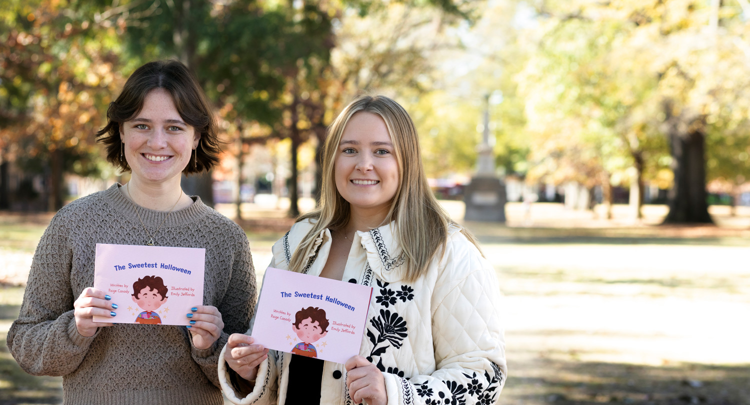 two women hold a book in an outdoor setting with trees in the background