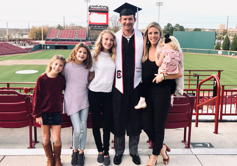 Adam Everett smiles at the baseball stadium with his family. He is wearing his graduation robe and mortarboard.