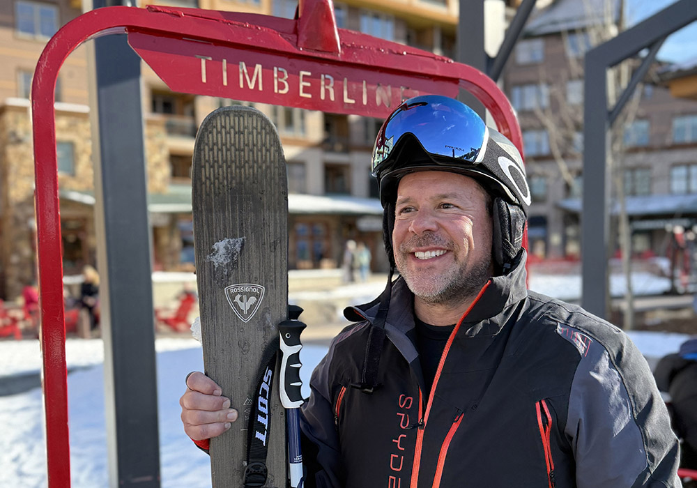 Man in ski helmet and goggles smiles while holding skis beneath a red Timberline sign at snowy mountain resort.