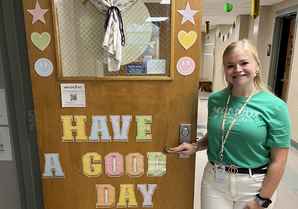 ophia Comose stands next to a decorated classroom door that reads, "Have A Good Day."