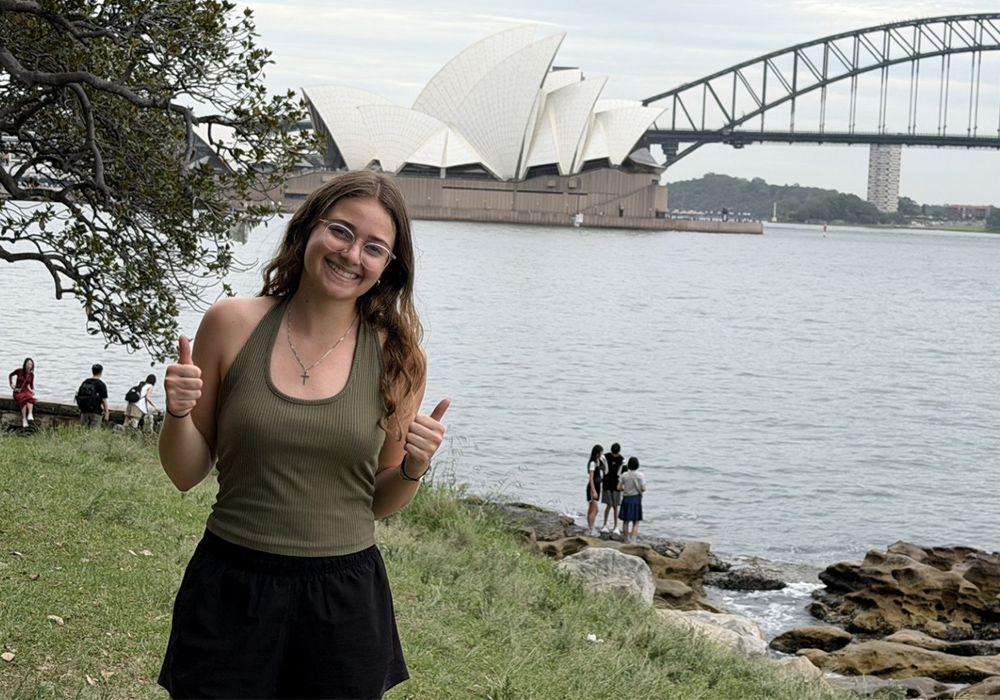 Ashley Chittum smiles and gives two thumbs up in Australia, with the Sydney Opera House in the background