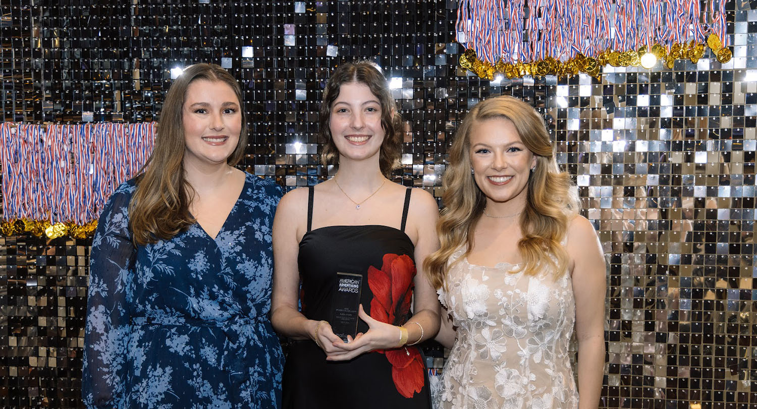 Three women in evening wear pose with trophy in festive setting