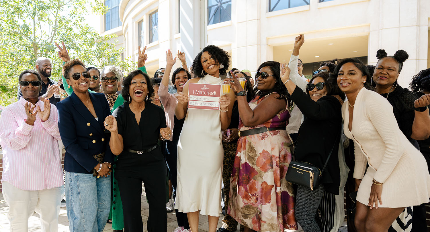 A group of people gathered outdoors celebrate as a woman in the center holds a sign reading “I Matched!” while smiling and raising her hand.